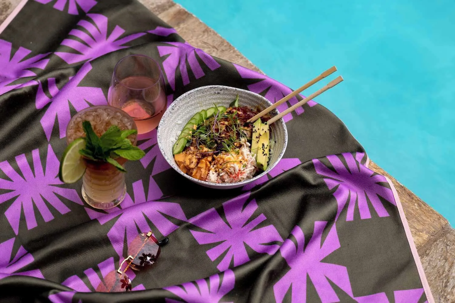 Poke bowl and drinks on a patterned towel beside the pool at Wayfinder Waikiki.
