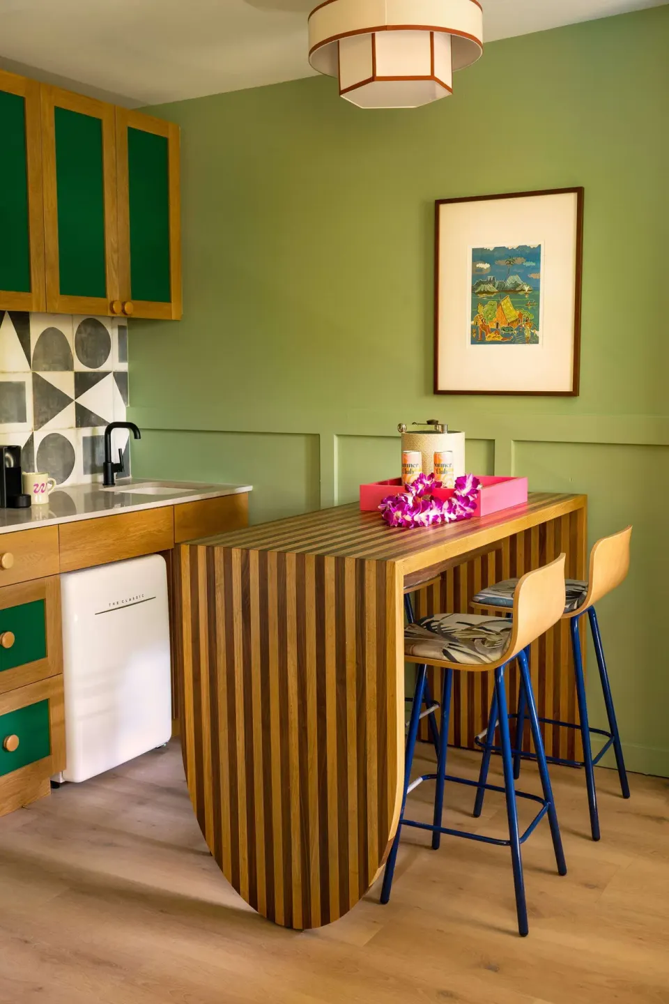 Studio kitchenette with striped bar, stools, sink, mini fridge, and green cabinets at Wayfinder Waikiki.