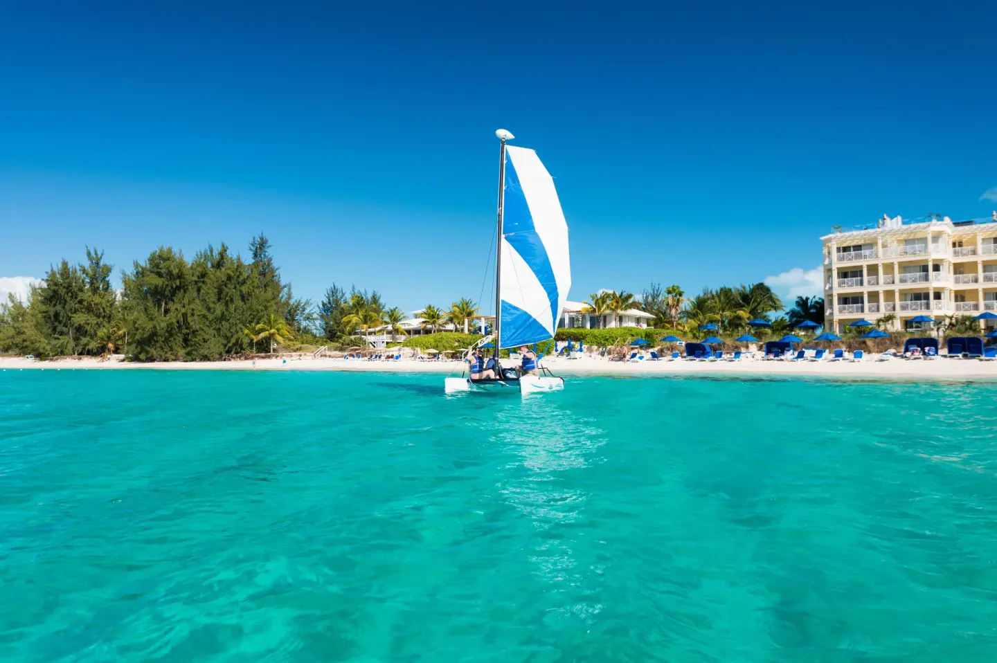 A small sailboat crosses turquoise water by the beach at Windsong on the Reef.
