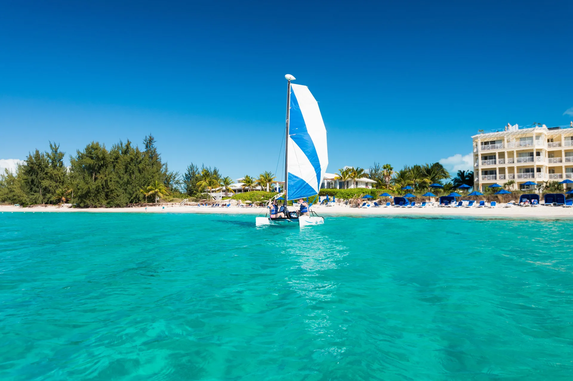 A small sailboat crosses turquoise water by the beach at Windsong on the Reef.