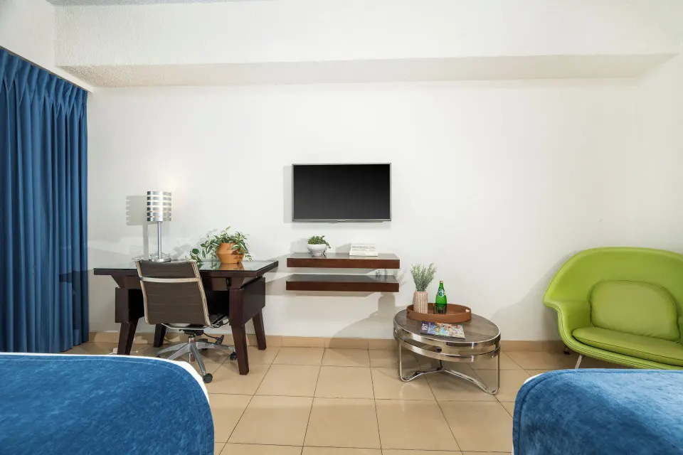Standard Room view of desk, wall TV, green chair, and bed corners at Windward Passage Hotel.