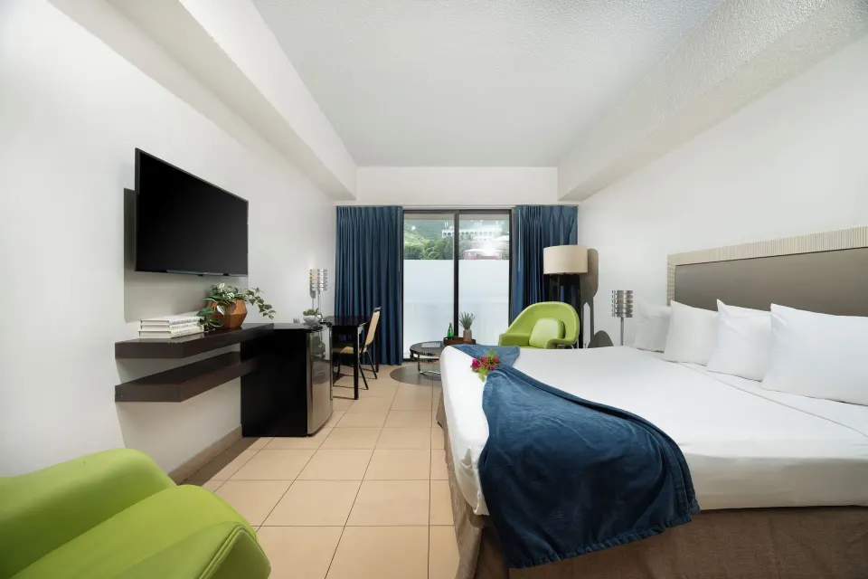Standard room with king bed, desk, TV and blue-curtained balcony doors at Windward Passage Hotel.