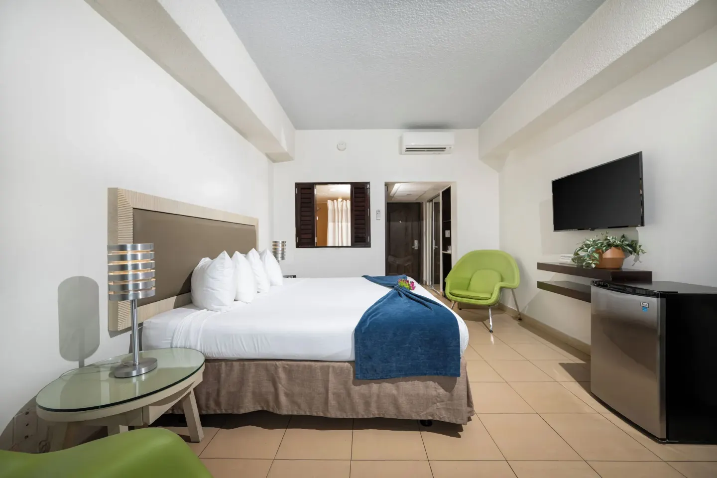 Standard room with king bed, wall TV, mini fridge and green chair at Windward Passage Hotel.
