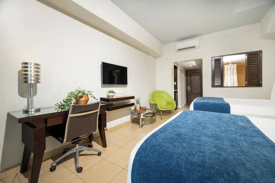 Standard Room with two beds, desk, TV, green chair, and interior window at Windward Passage Hotel.