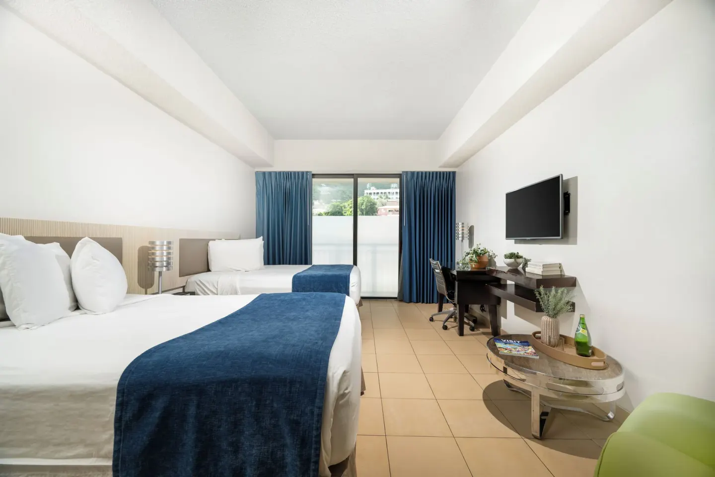 Standard Room with two beds, blue curtains, desk, TV, and balcony doors at Windward Passage Hotel.